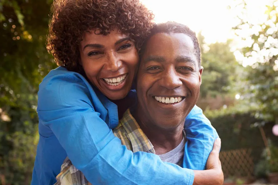 Autoimmune-Doctor A middle-aged African American couple outside, the woman embracing the man from behind pressing her cheek to his, both are smiling due to successful Autoimmune Disorder treatment from Dr. Stephanie King in Salt Lake City.