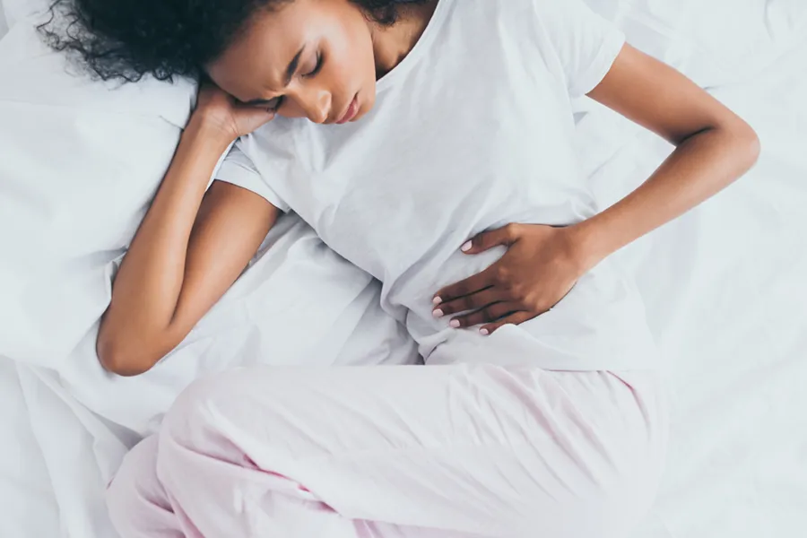 Crohns-Disease-Clinic A dark-skinned woman in white clothes lays on her bed clutching her middle before getting treatment for Crohn's Disease from Dr. Stephanie King in Salt Lake City.