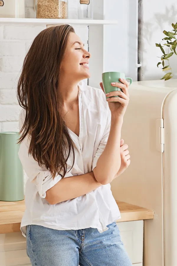 Crohns-Disease-Treatment A woman with a white blouse an jeans drinking tea in her kitchen, smiling after getting treatment for Crohn's Disease from Dr. Stephanie King in Salt Lake City.