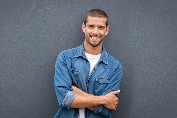 Hormones-for-Men-Doctor A man in a denim shirt stands smiling against a gray-blue wall, pleased with his testosterone hormone therapy from Dr. Stephanie King in Salt Lake City.