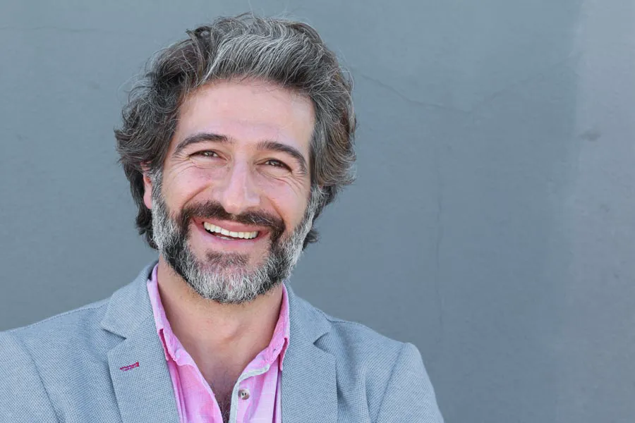 Hypogonadism-Clinic A smiling man with salt and pepper hair and beard wearing a pink shirt and gray blazer stands agains a gray wall. Get hypogonadism treatment from Dr. Stephanie King in Salt Lake City.