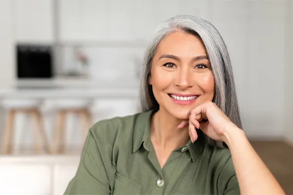 Menopause-Clinic An asian woman with long gray hair and a green shirt sitting and smiling on her couch, happy with her menopause care from [NAME} in Salt Lake City.