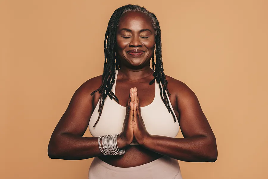 Obesity-Clinic An older African American woman confidently posing against a warm background smiling with hands in a prayer pose. Dr. Stephanie King treats obesity using functional medicine in Salt Lake City.