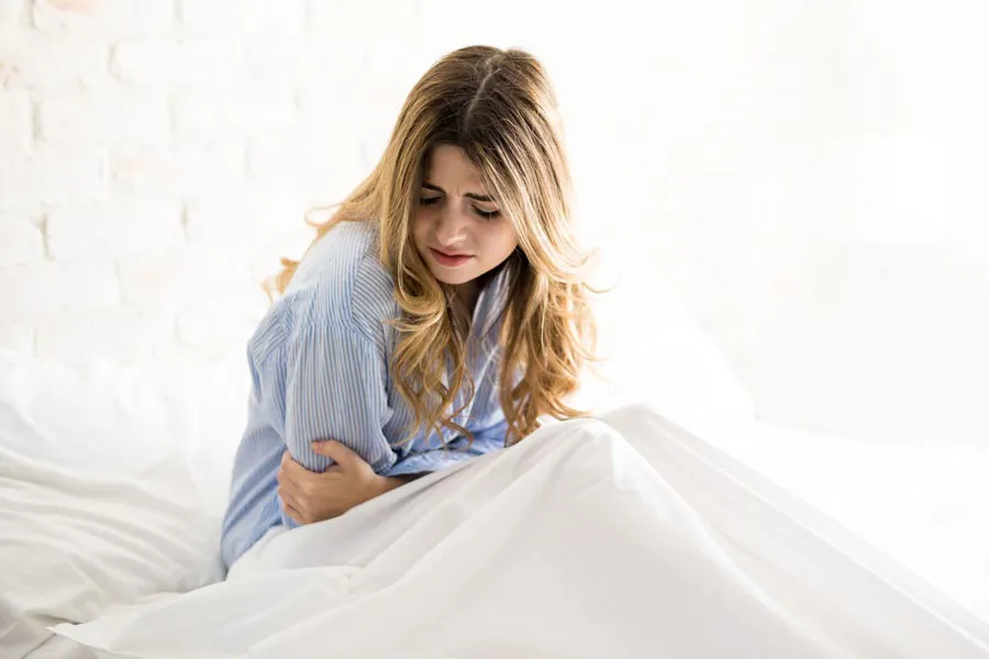 PMS-Therapy A woman with long blonde hair sits in her bed holding her stomach in pain, considering getting care for PMS from Dr. Stephanie King in Salt Lake City.