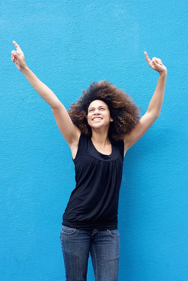 PMS-Treatment A woman in a dark blue tank top standing in front of a bright blue wall, raising her arms in celebration of relief from PMS from Dr. Stephanie King in Salt Lake City.