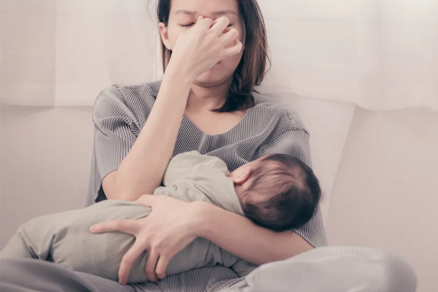 Postpartum-Hormone-Imbalance-Clinic A woman nursing an infant on couch rubs her eyes with exhaustion. Get treatment for postpartum hormone imbalance from Dr. Stephanie King in Salt Lake City.