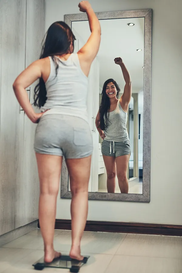 Tirzepatide-Treatment A woman in workout clothes stands on a scale looking in the mirror and pumping her hands in celebration of her successful Tirzepatide treatment from Dr. Stephanie King in Salt Lake City.